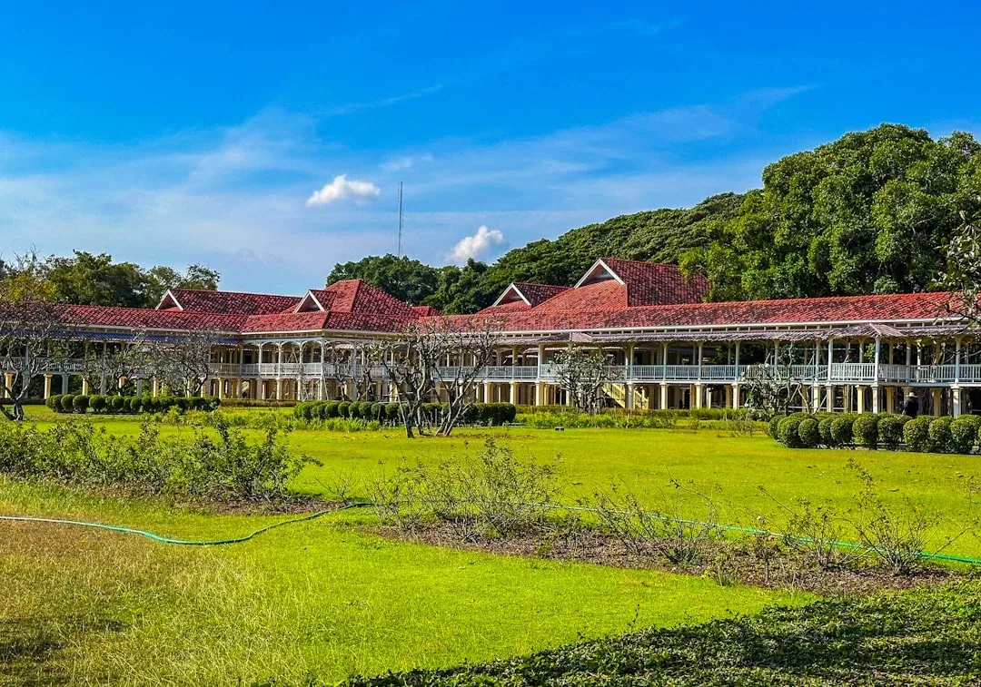 A grand colonial-style building with a red roof and white verandas on a green lawn in Phetchaburi, Thailand.