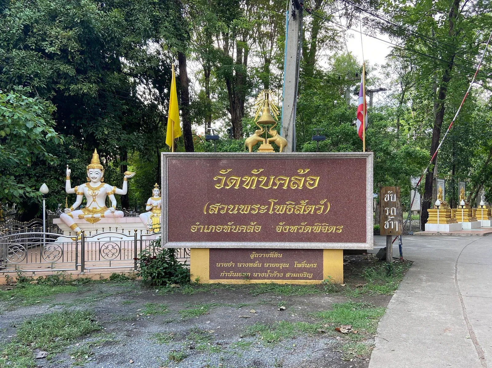 Entrance to Wat Thap Khlo in Phichit Province, Thailand, with a large brown sign, a white multi-armed deity statue, and green trees.