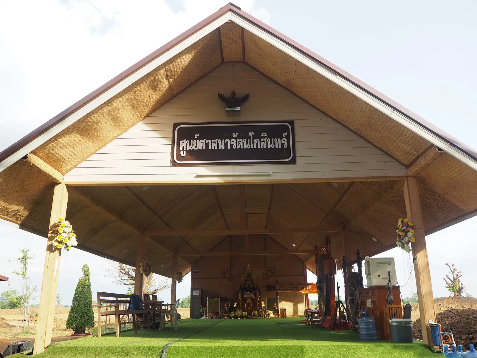 A modern, open-air pavilion with a gabled roof and a Thai sign, housing a Buddhist altar with statues and offerings on a green turf floor.