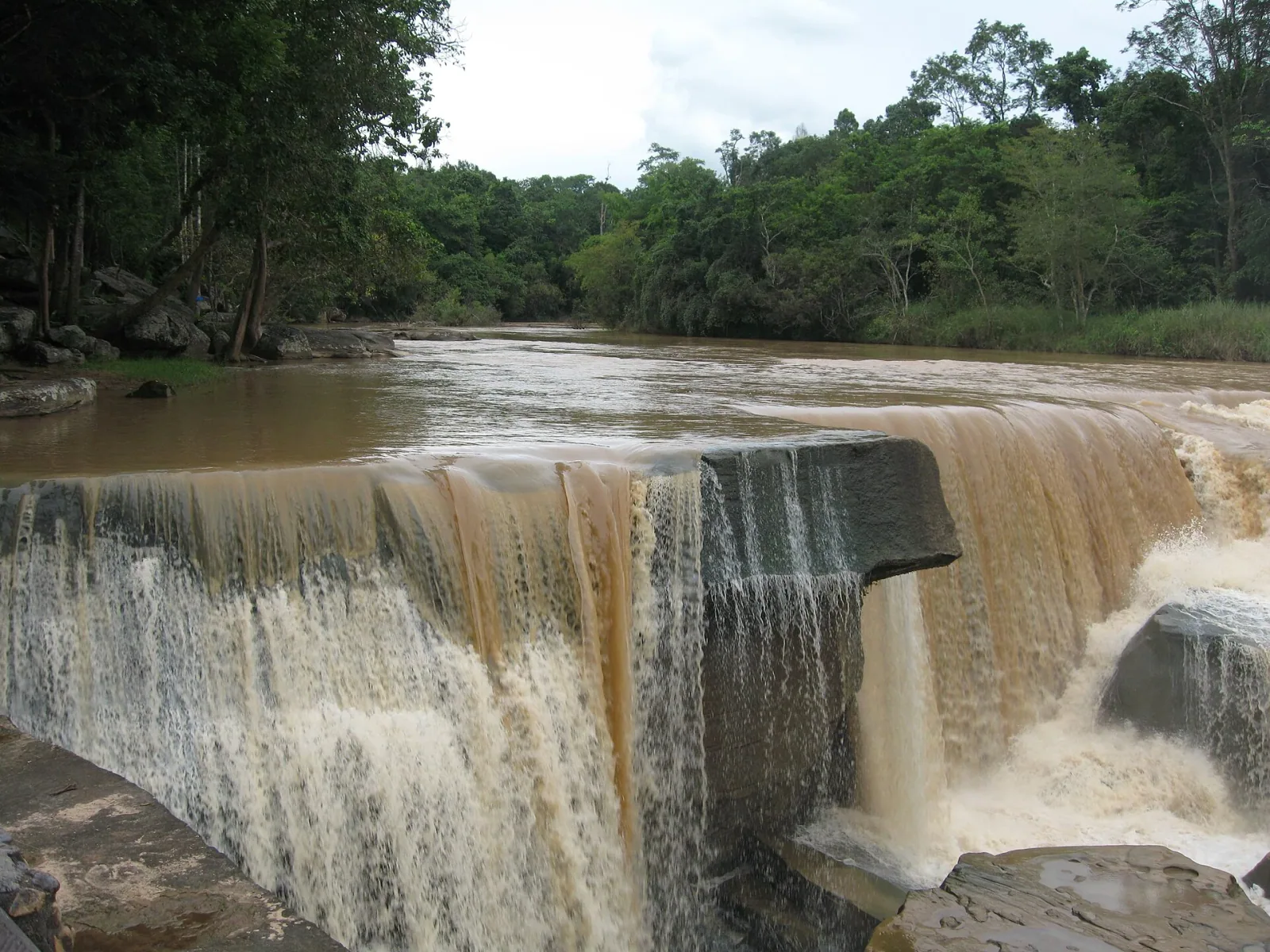 Kaeng Sopha Waterfall in Phitsanulok, Thailand, showing a wide, muddy river cascading over natural rock formations amidst dense green forest.