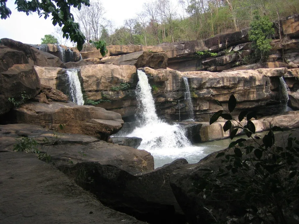 Kaeng Sopha Waterfall in Phitsanulok, Thailand, showing multiple streams of water flowing over wide, layered rock formations into a pool below, with trees in the background.