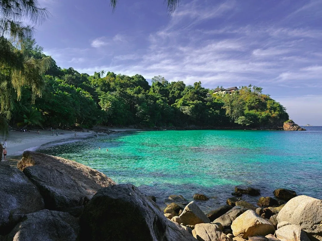 Scenic view of Banana Beach in Phuket, Thailand, with clear turquoise water, a sandy shore, and lush green hills.