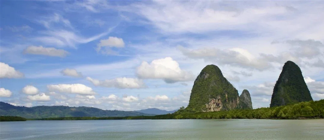 Panoramic view of green, tree-covered limestone karsts rising from calm waters in Phang Nga Bay, Thailand, under a blue sky with white clouds.