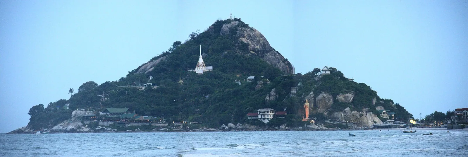 Panoramic view of Khao Takiap, a large green and rocky hill rising from the sea, featuring a white pagoda and a golden Buddha statue on its slopes, with small boats in the foreground under a light blue sky.