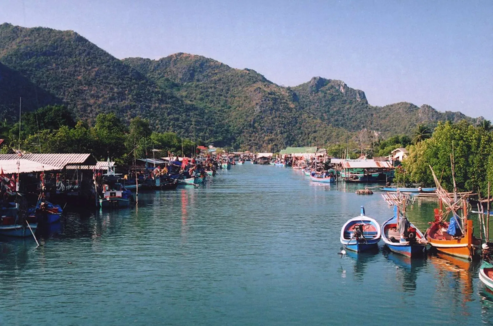 Traditional Thai fishing boats docked along a waterway, with stilted buildings on the banks and forested mountains in the background.