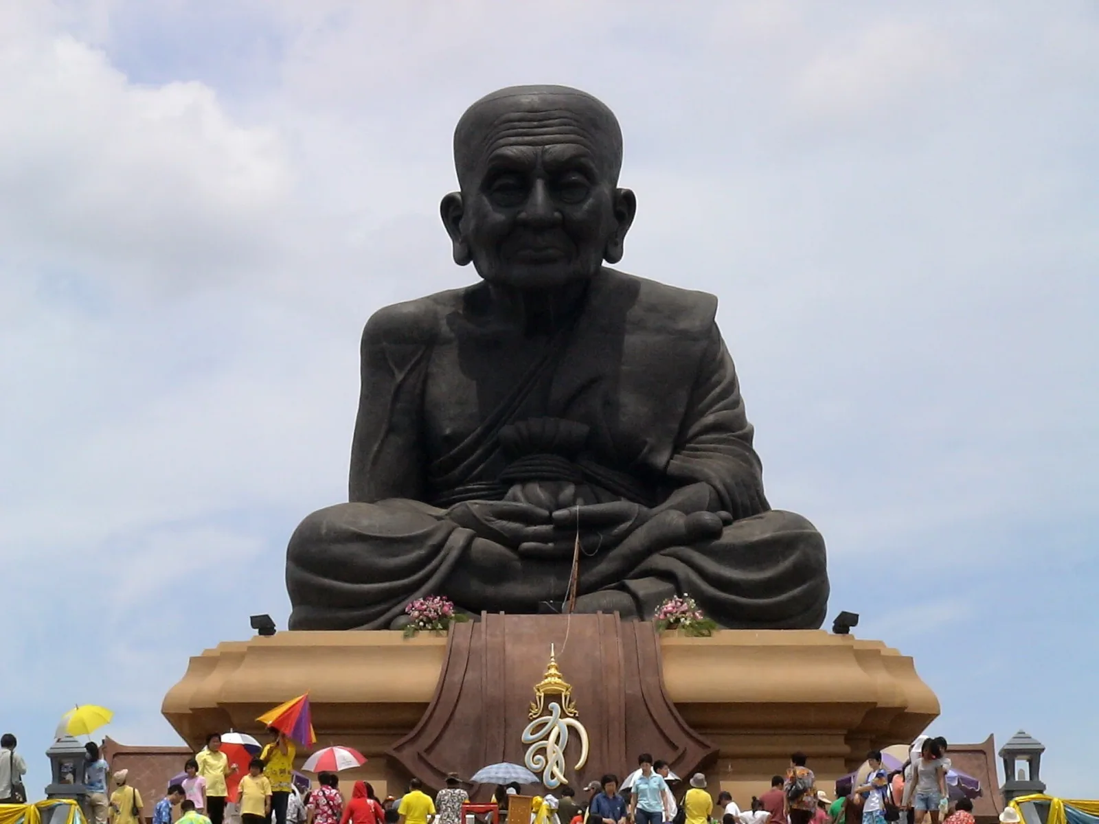 A massive, dark statue of a revered Buddhist monk, Luang Pu Thuat, seated in meditation, with a crowd of visitors gathered at its ornate base under a partly cloudy sky.
