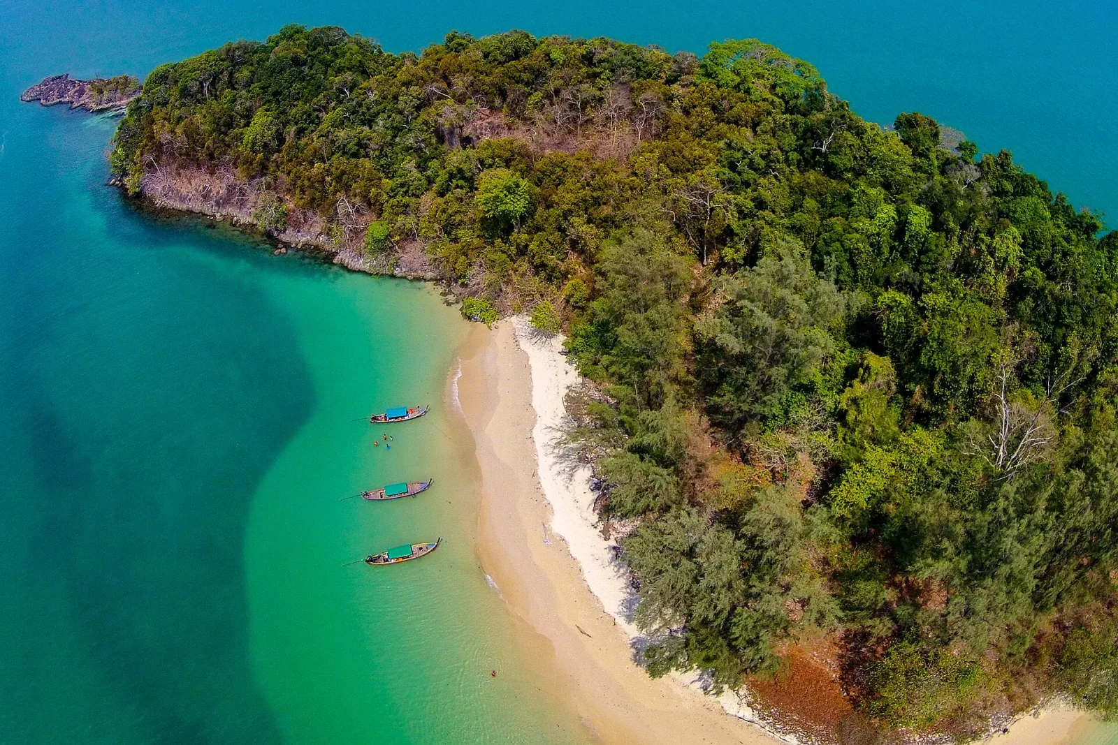 Aerial view of a tropical island with dense green foliage, a white sand beach, and three longtail boats floating in clear turquoise water.
