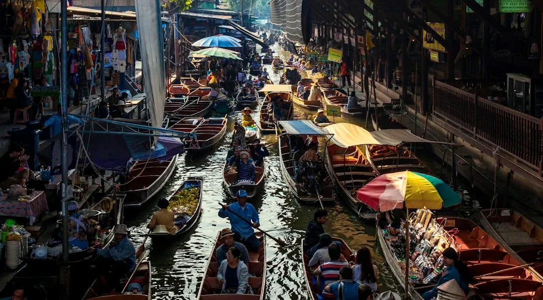 Overhead view of Damnoen Saduak Floating Market in Ratchaburi, Thailand, showing many wooden boats with vendors and tourists on a busy canal, surrounded by market stalls.