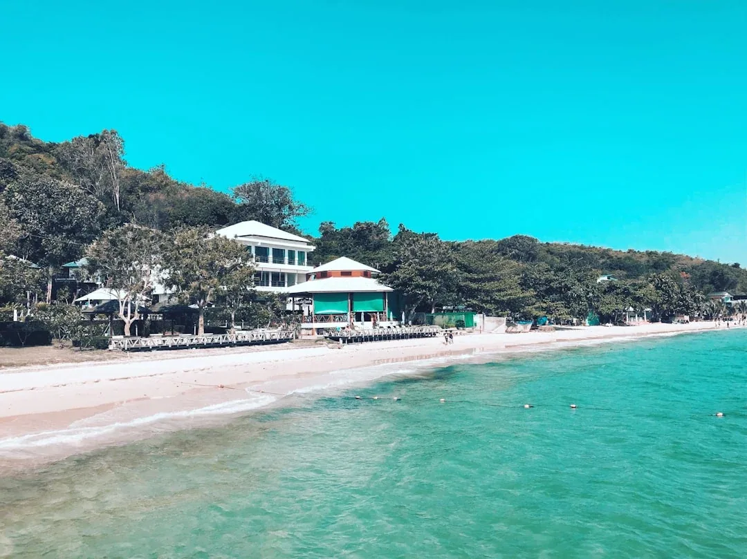 A white multi-story building and a green-roofed pavilion stand on a sandy beach next to turquoise ocean water, with a tree-covered hill in the background on Ko Samet Island, Rayong.