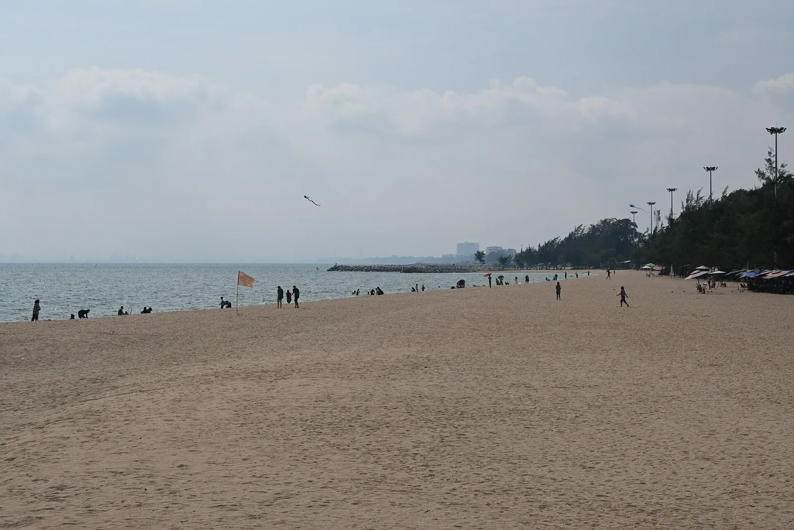 A wide, hazy view of a sandy beach with a calm sea, a few distant people, and a tree-lined area with light poles on the right.