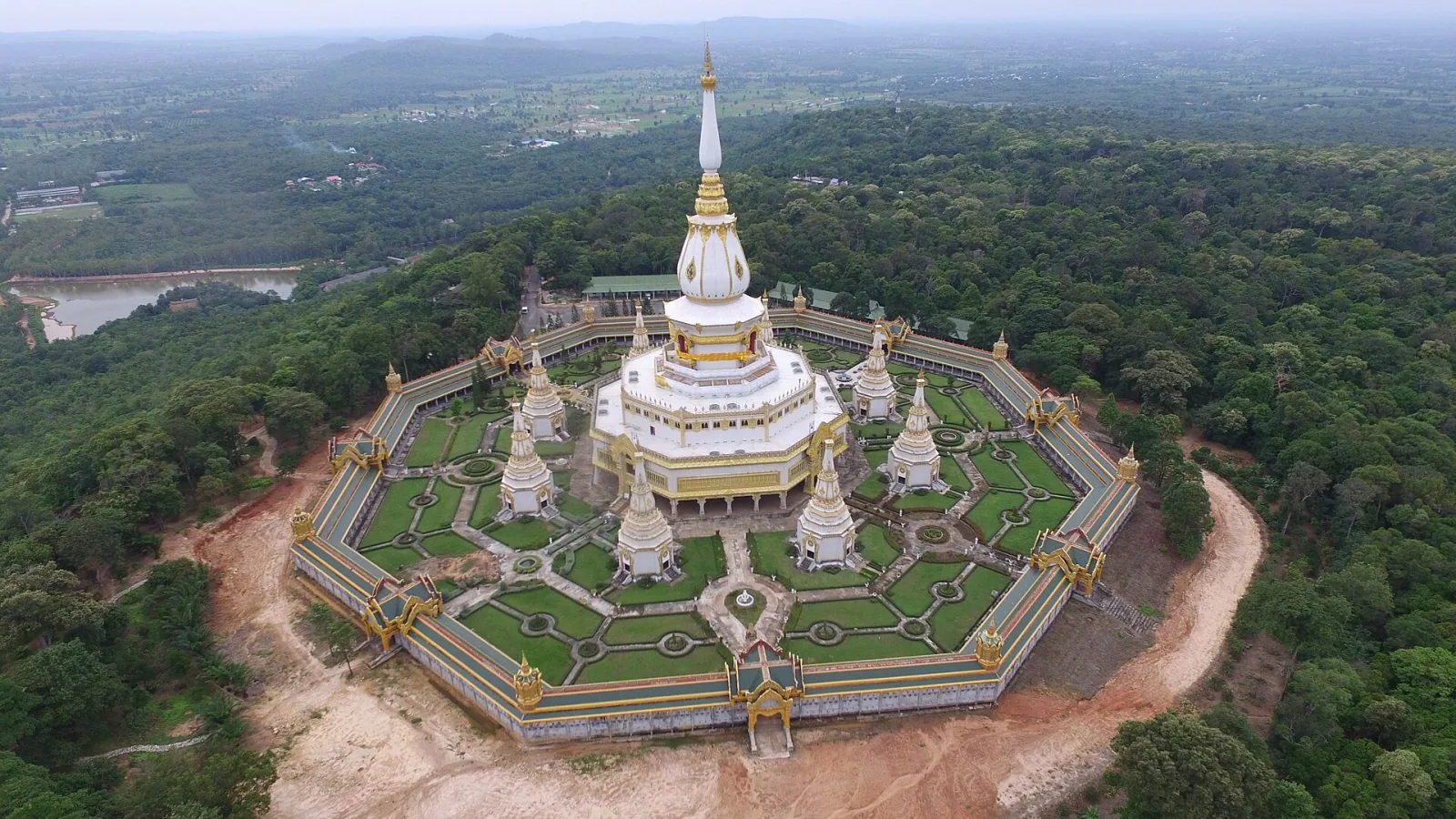 Aerial view of Phra Maha Chedi Chai Mongkol, a large white and gold multi-tiered chedi complex with surrounding gardens, atop a forested hill in Roi Et province.