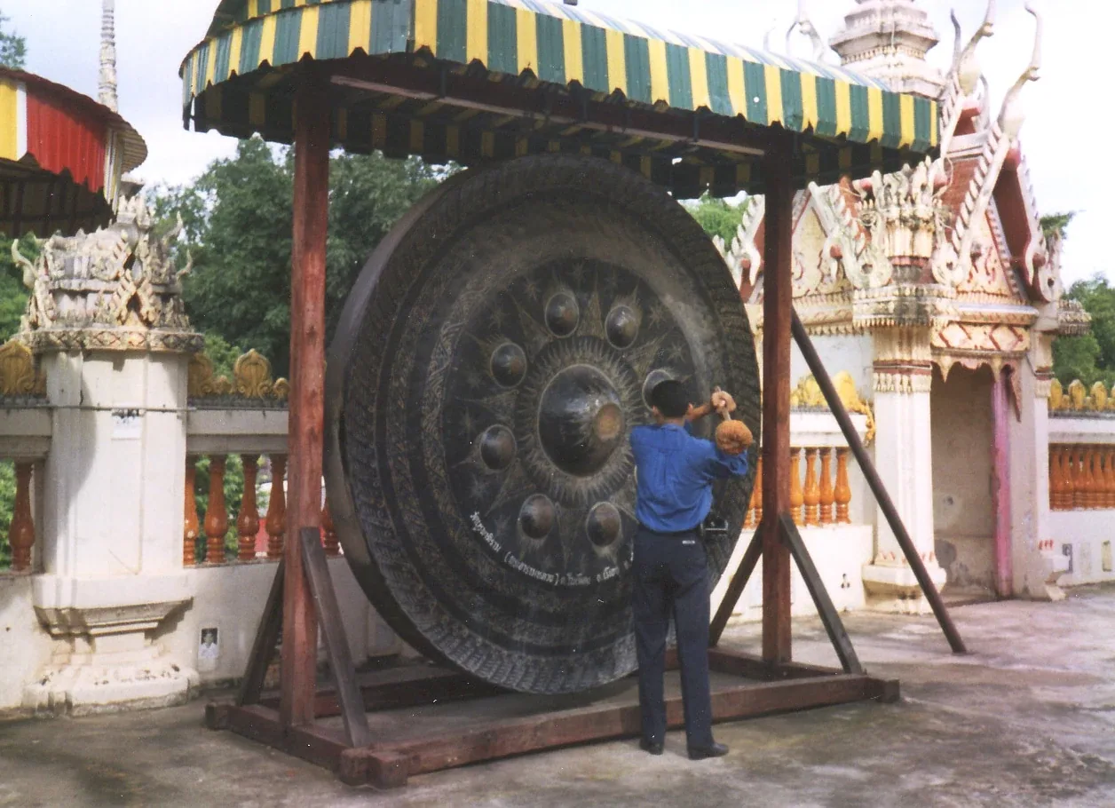 A person in a blue shirt strikes a massive, ornate bronze gong under a yellow and green striped canopy at a Thai temple.