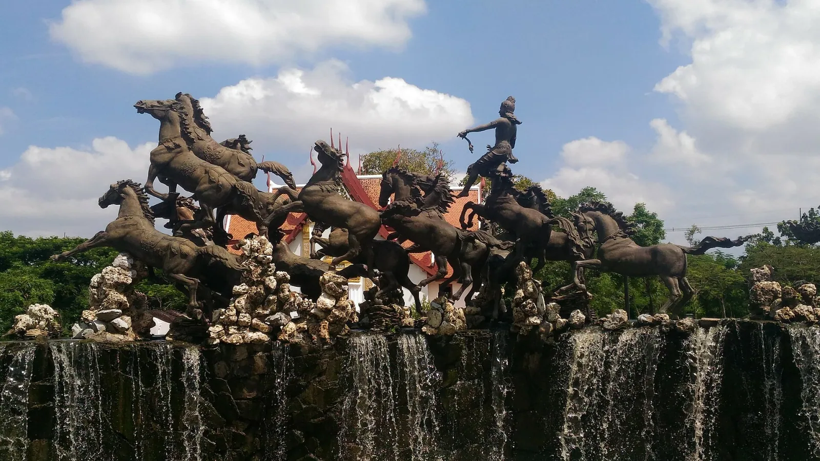A bronze sculpture of a deity, possibly Surya, standing on a chariot pulled by many horses, with water cascading from its base, set against a blue sky with clouds and green foliage.
