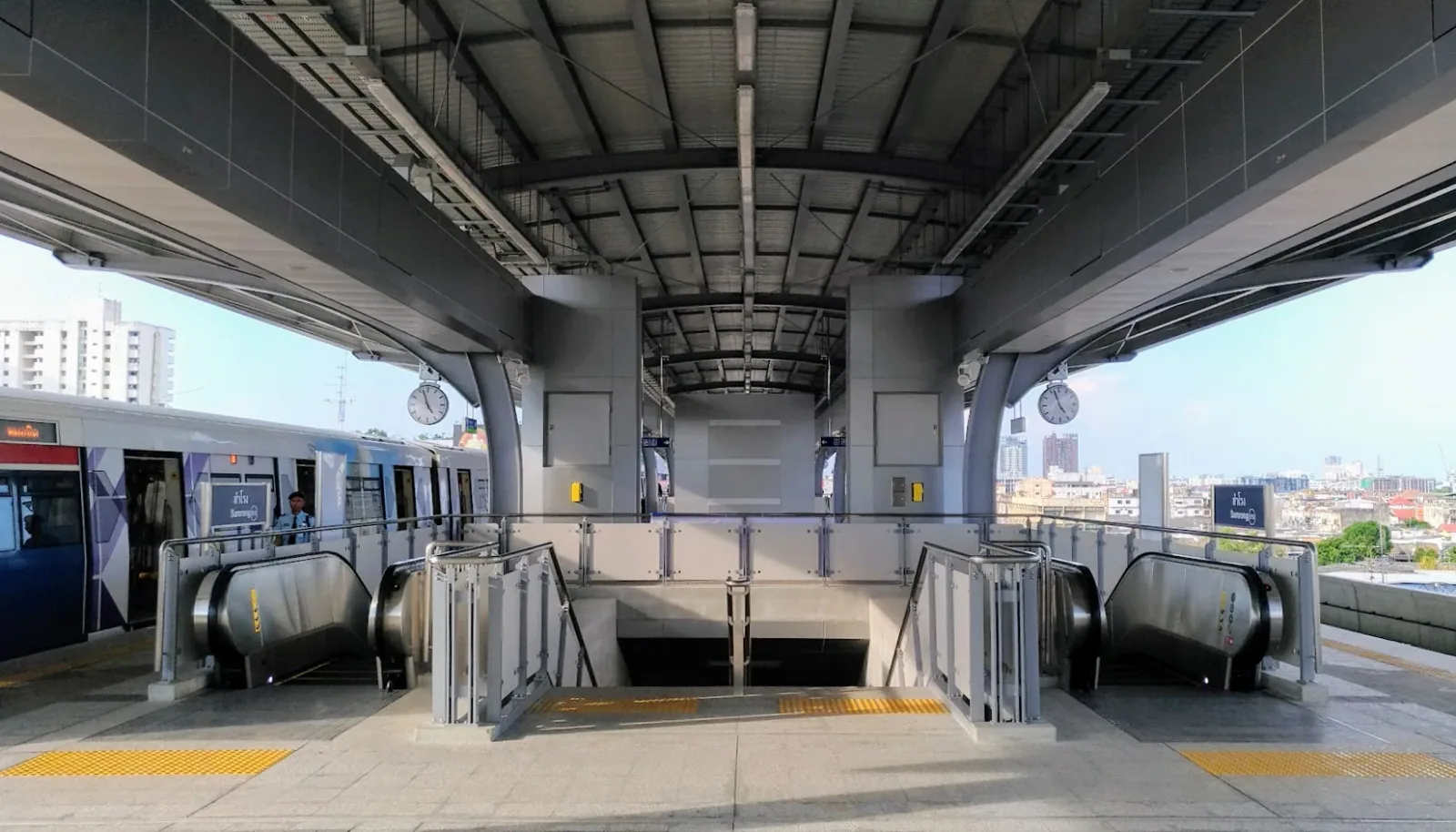 Elevated view of the modern BTS Samrong Skytrain station platform with a train stopped, escalators, and a city skyline in the background.