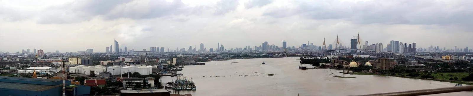 Panoramic view of the Chao Phraya River in Samut Prakan, showing industrial docks, a distant city skyline, and the distinctive Bhumibol Bridge with its two cable-stayed towers.