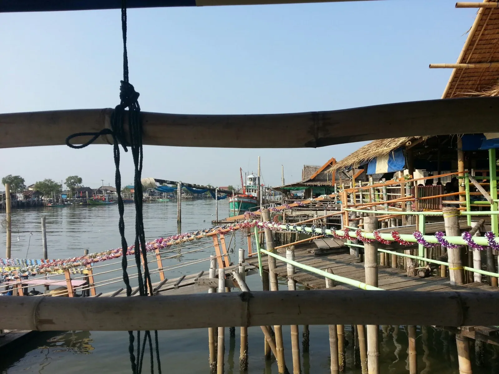 Stilt houses of a fishing village line a river in Samut Sakhon, Thailand, with fishing boats visible in the distance.