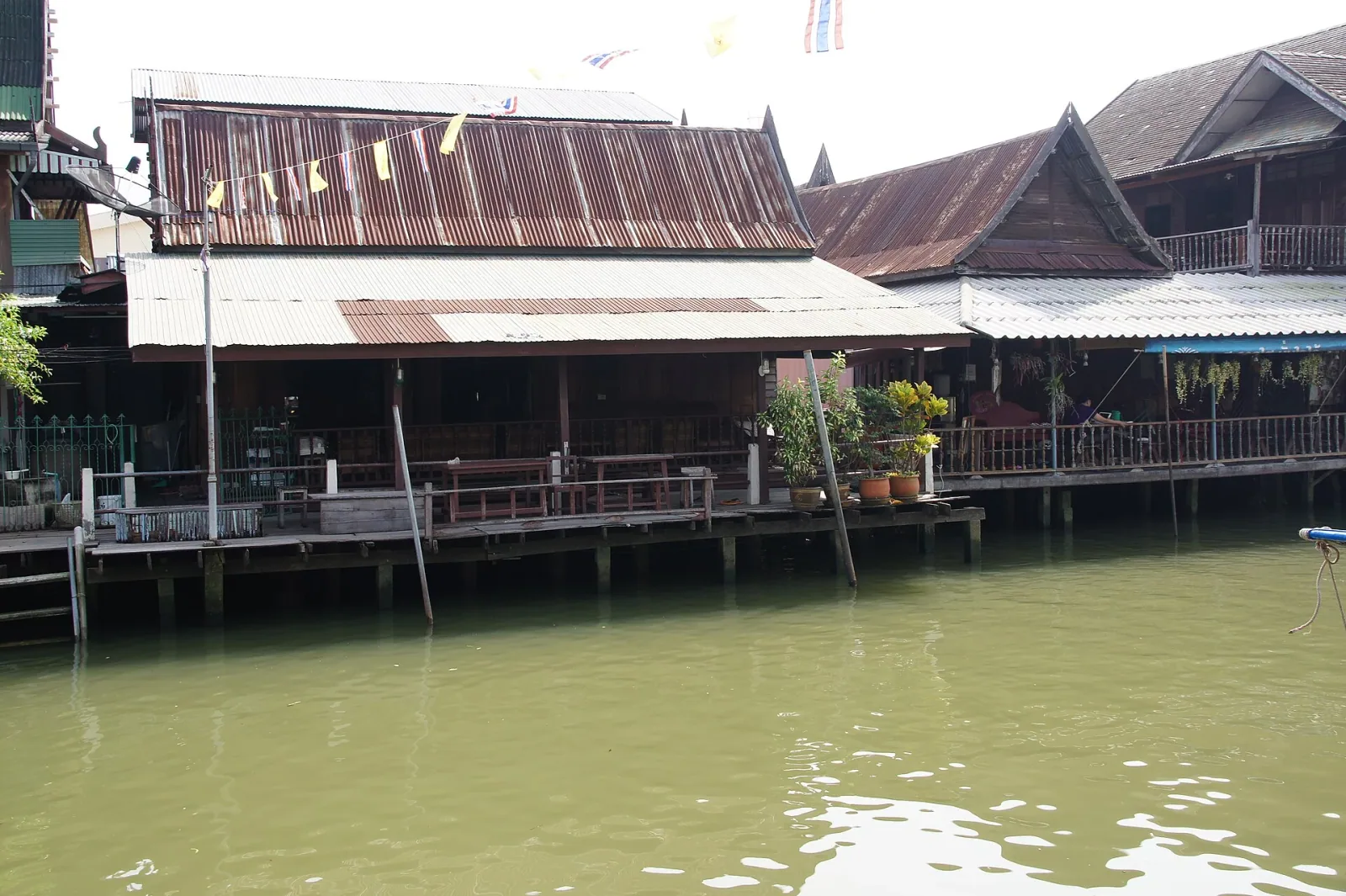 Traditional wooden stilt houses with corrugated roofs line a green canal in Amphawa, Samut Songkhram, Thailand.