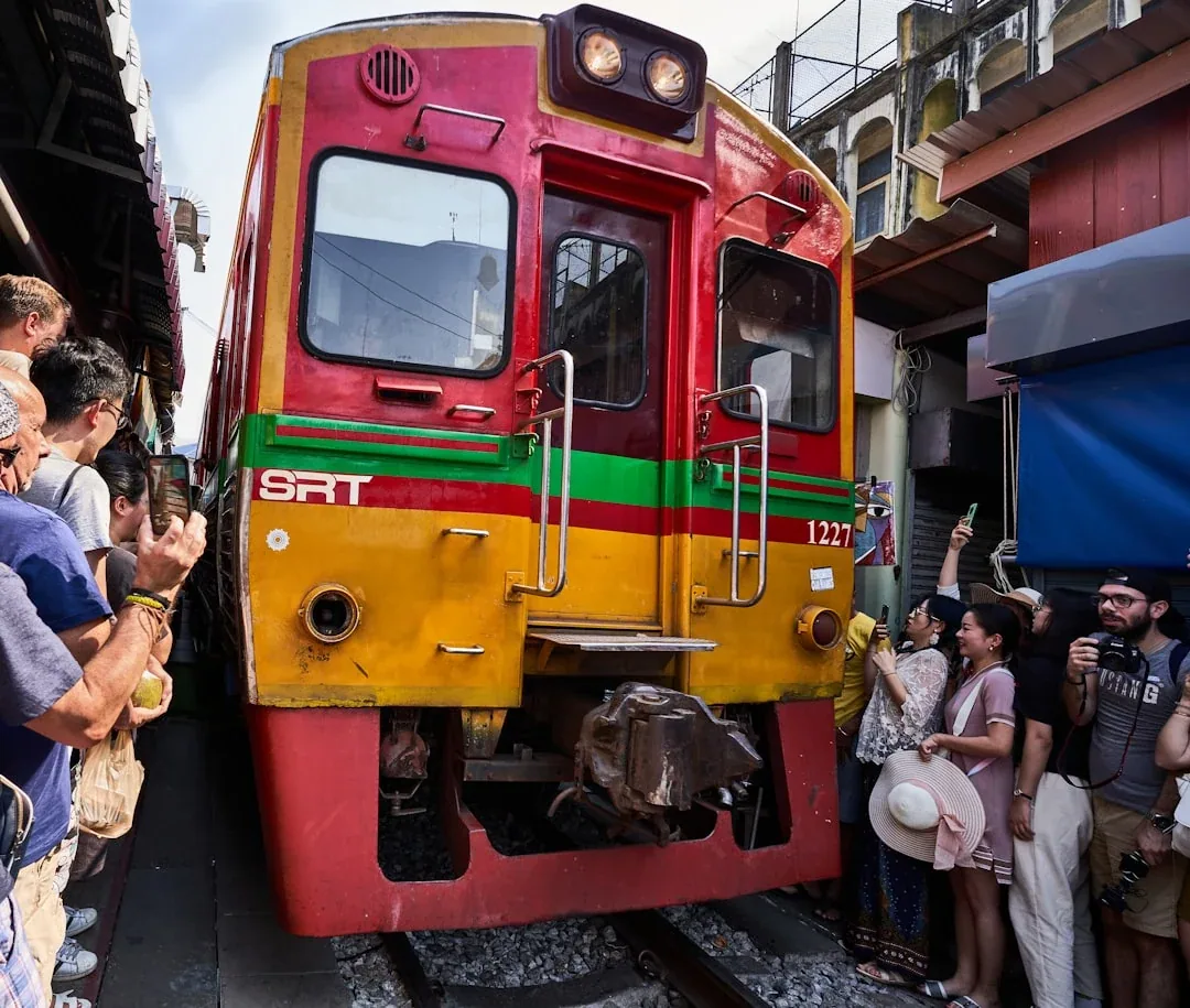 A red, yellow, and green train with 'SRT' on its front passes through the narrow Maeklong Railway Market, surrounded by tourists taking pictures.