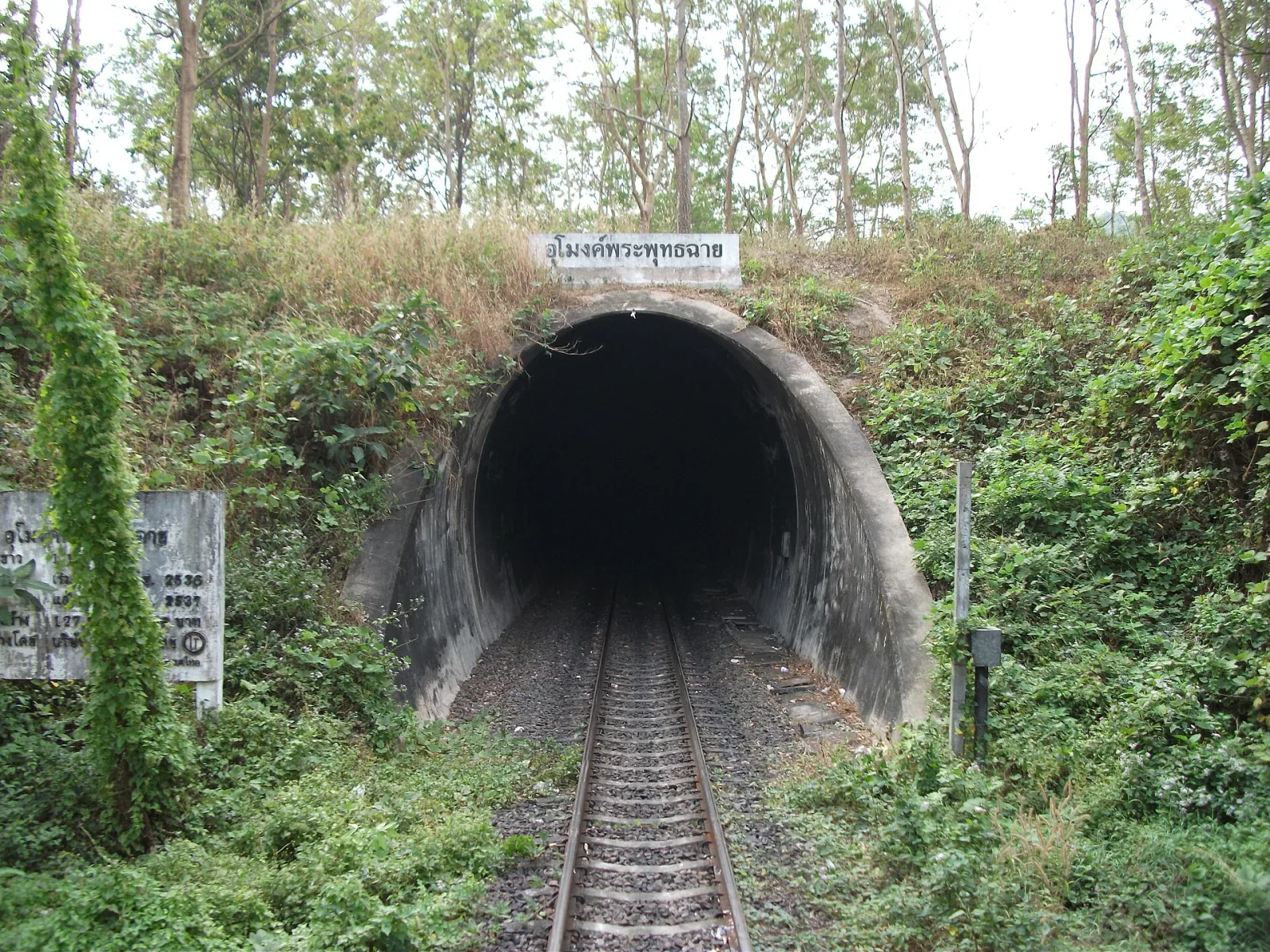 The entrance to Phra Phutthachai Tunnel, a railway tunnel in Saraburi, Thailand, surrounded by lush green trees and foliage, with train tracks leading into its dark opening.