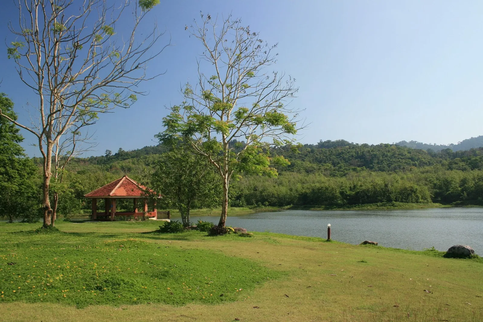 A tranquil lake with a red-roofed pavilion on a grassy shore, backed by dense green forests and hills under a bright blue sky.