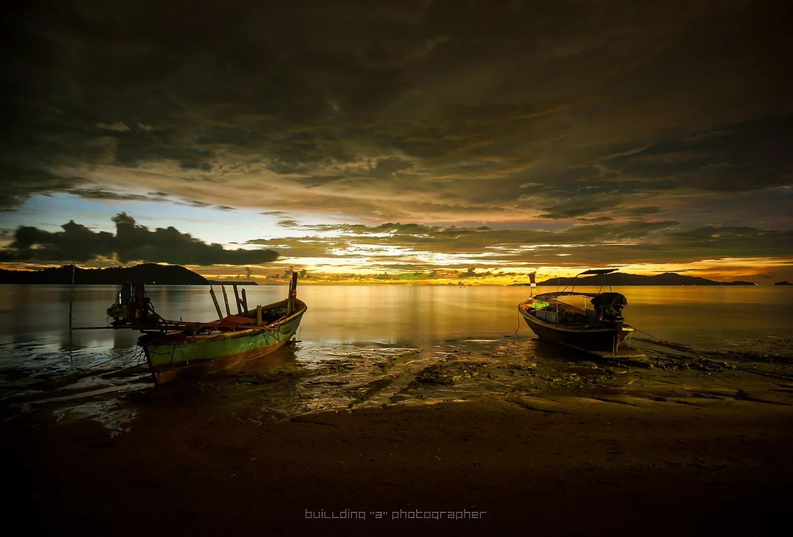 Two traditional longtail boats sit on a beach at sunset, with a vibrant orange and yellow sky reflecting on the calm water and distant islands.