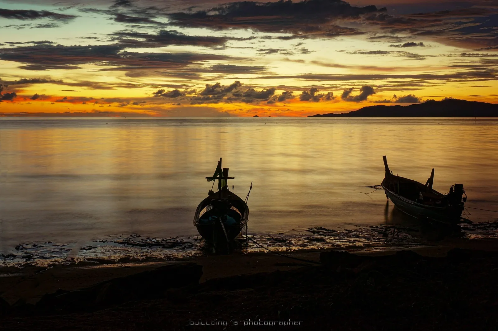 Two traditional Thai longtail boats are silhouetted on a beach at sunset, with the sky ablaze in orange and yellow over calm ocean waters and distant islands.