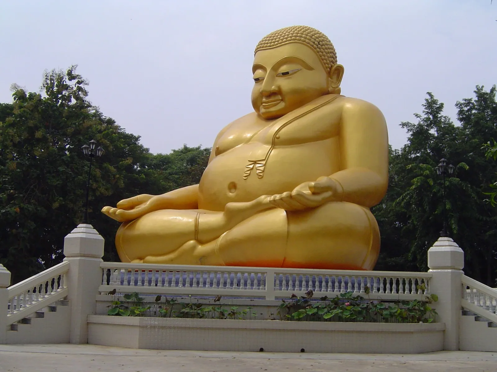 A large, golden statue of Phra Sangkatchai, a plump Buddhist monk, sits cross-legged on a white tiered platform with railings, surrounded by green trees.