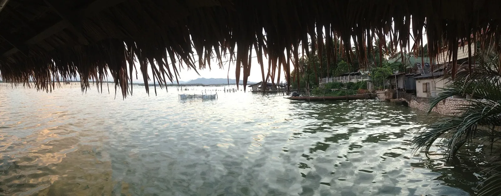 Panoramic view from under a dark thatched roof across a calm body of water, with stilt houses and trees on the right bank and distant hills.