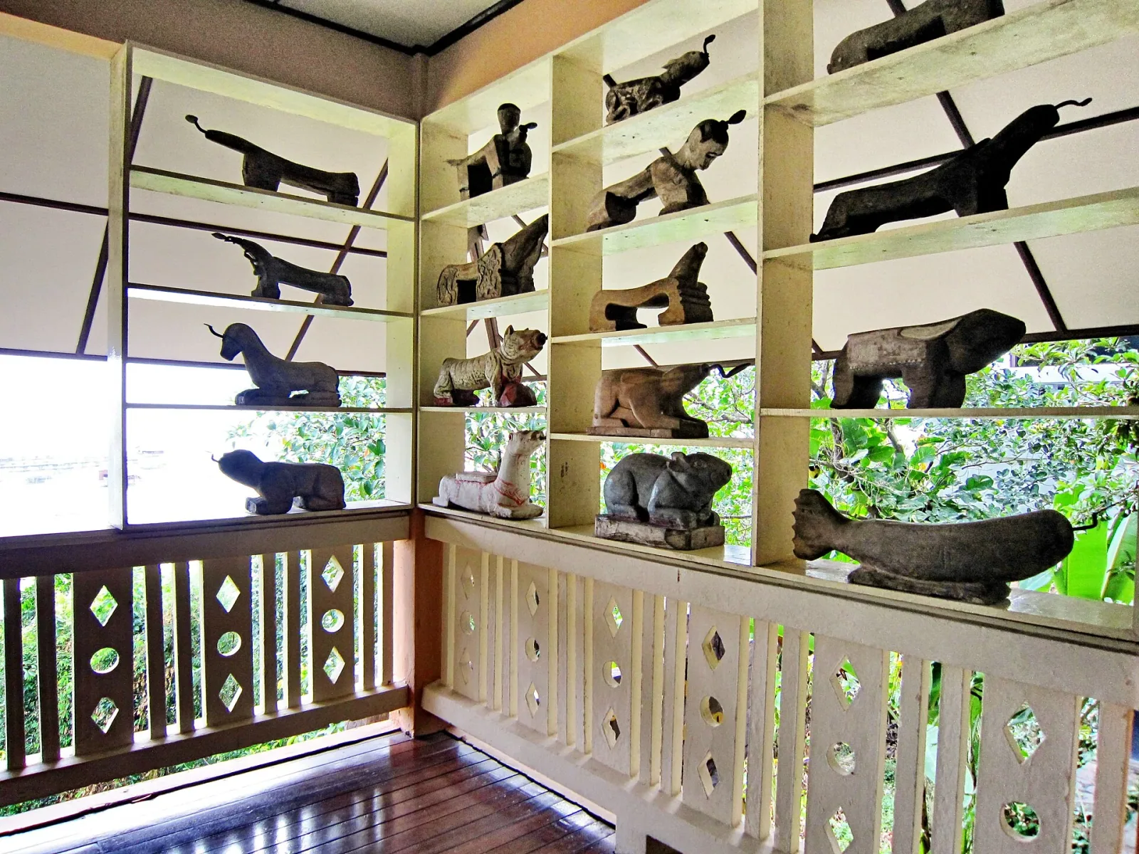 A display of traditional wooden and stone sculptures, including various animal figures, on white shelves inside the Institute for Southern Thai Studies, with a balcony railing and green foliage visible.
