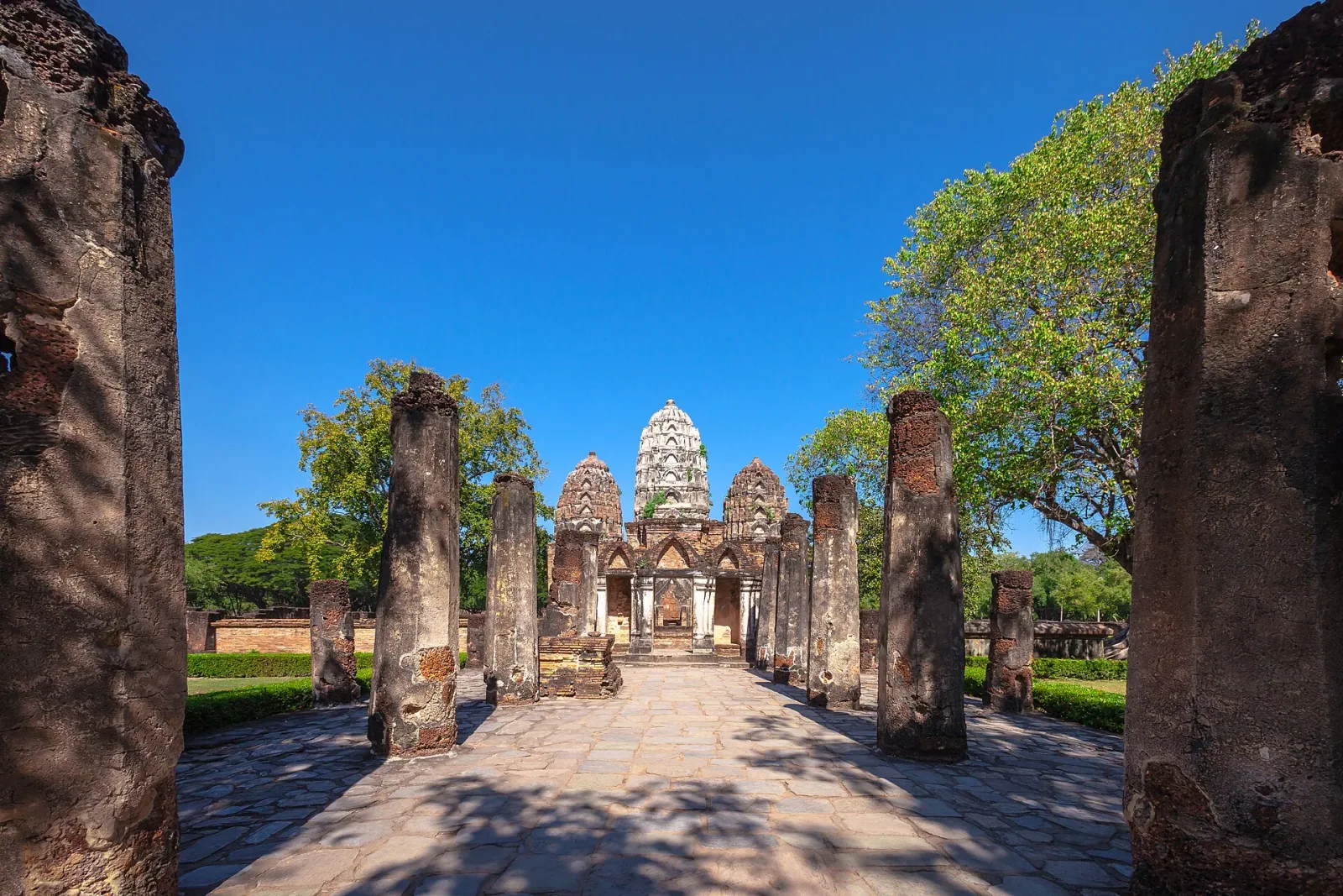 The ancient Wat Sri Sawai temple in Sukhothai, Thailand, featuring a central white Khmer-style prang and two smaller prangs, viewed from a stone pathway lined with weathered brick columns under a bright blue sky.