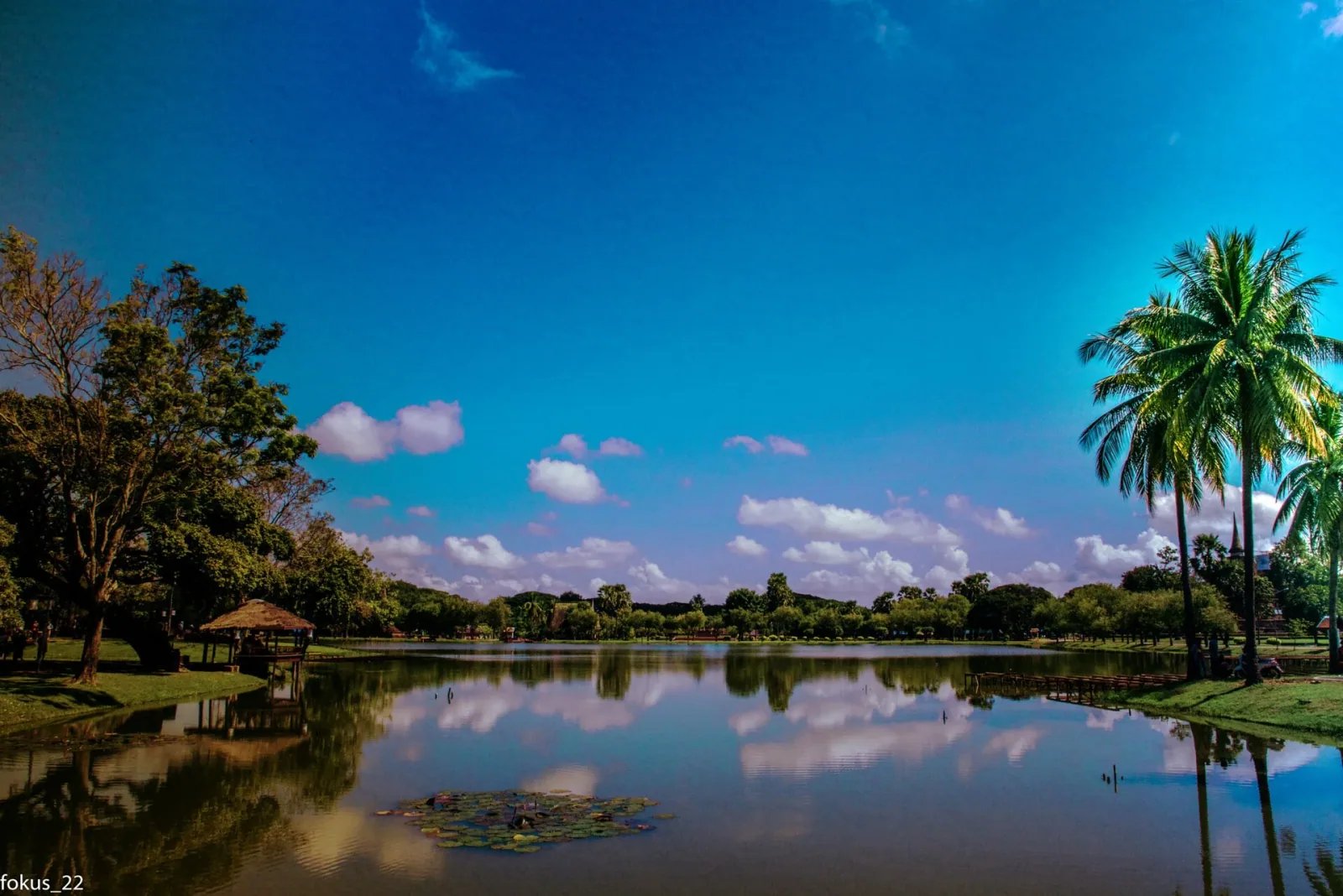 A serene lake reflects a bright blue sky with white clouds, bordered by green trees and palm trees, with a small thatched pavilion on the left shore.