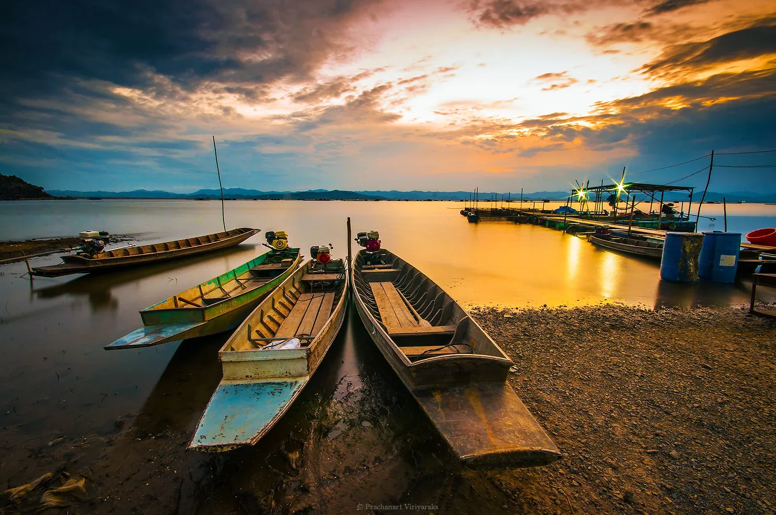 Longtail boats rest on the muddy shore of Kraseao Dam Reservoir at sunset, with a long wooden pier stretching into the water reflecting the golden sky.
