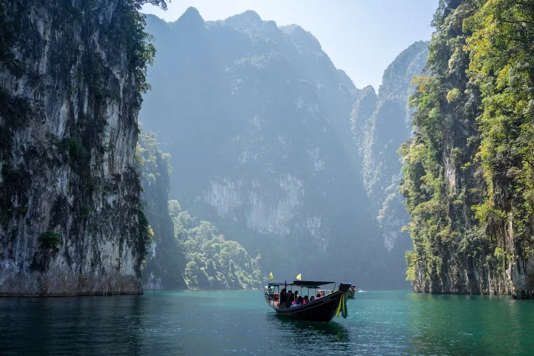 A traditional longtail boat with passengers on the emerald waters of Cheow Lan Lake, framed by steep, jungle-covered limestone cliffs in Khao Sok National Park, Surat Thani, Thailand.