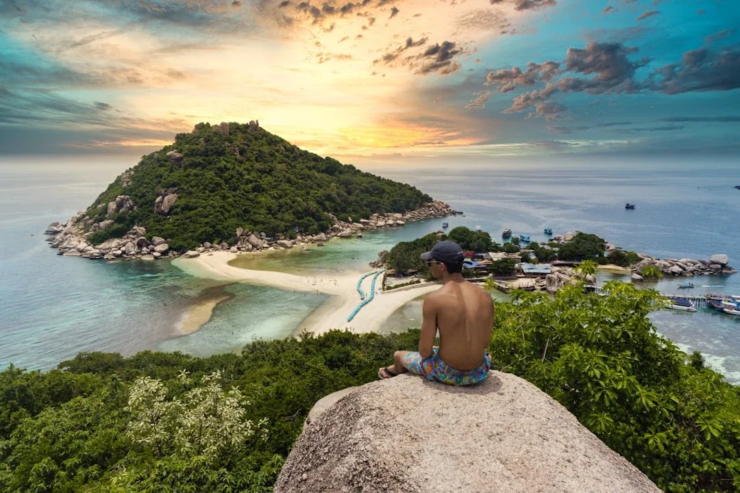 Panoramic view of Koh Nang Yuan island at sunset, showing its three small landmasses connected by white sandbars, surrounded by clear turquoise water. A person sits on a rock in the foreground, looking out at the scene.