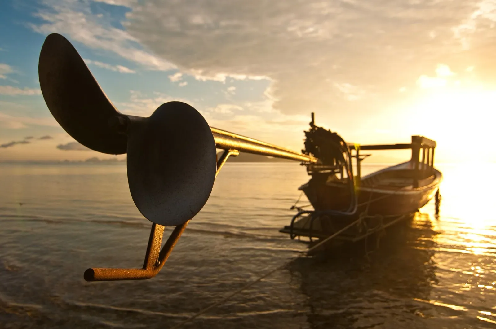 Close-up of a long-tailed boat's propeller in the foreground, with the boat and a golden sunrise or sunset over the ocean in the background.