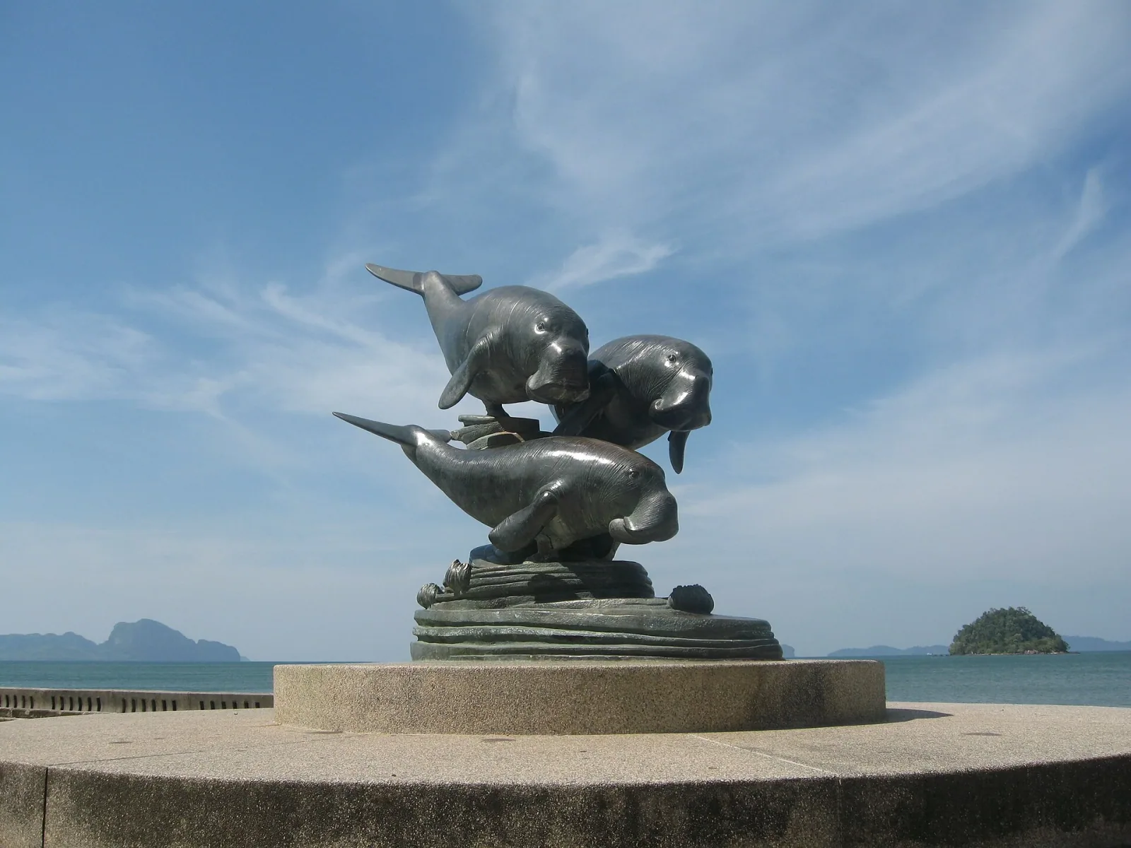 Bronze statue of three dugongs on a concrete pedestal at Pak Meng Beach, with the ocean and distant islands under a blue sky.