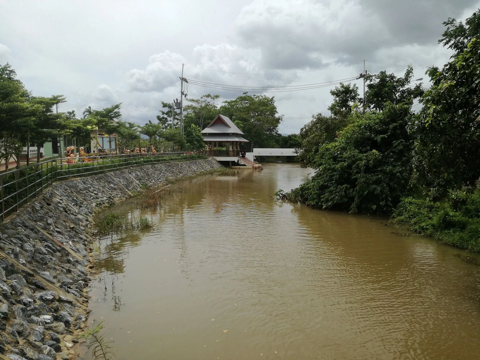 The Nam Sai river, a murky brown waterway, flows past a stone-lined bank with a metal fence on the left and dense green foliage on the right. A traditional wooden pavilion on stilts overlooks the river under a cloudy sky.