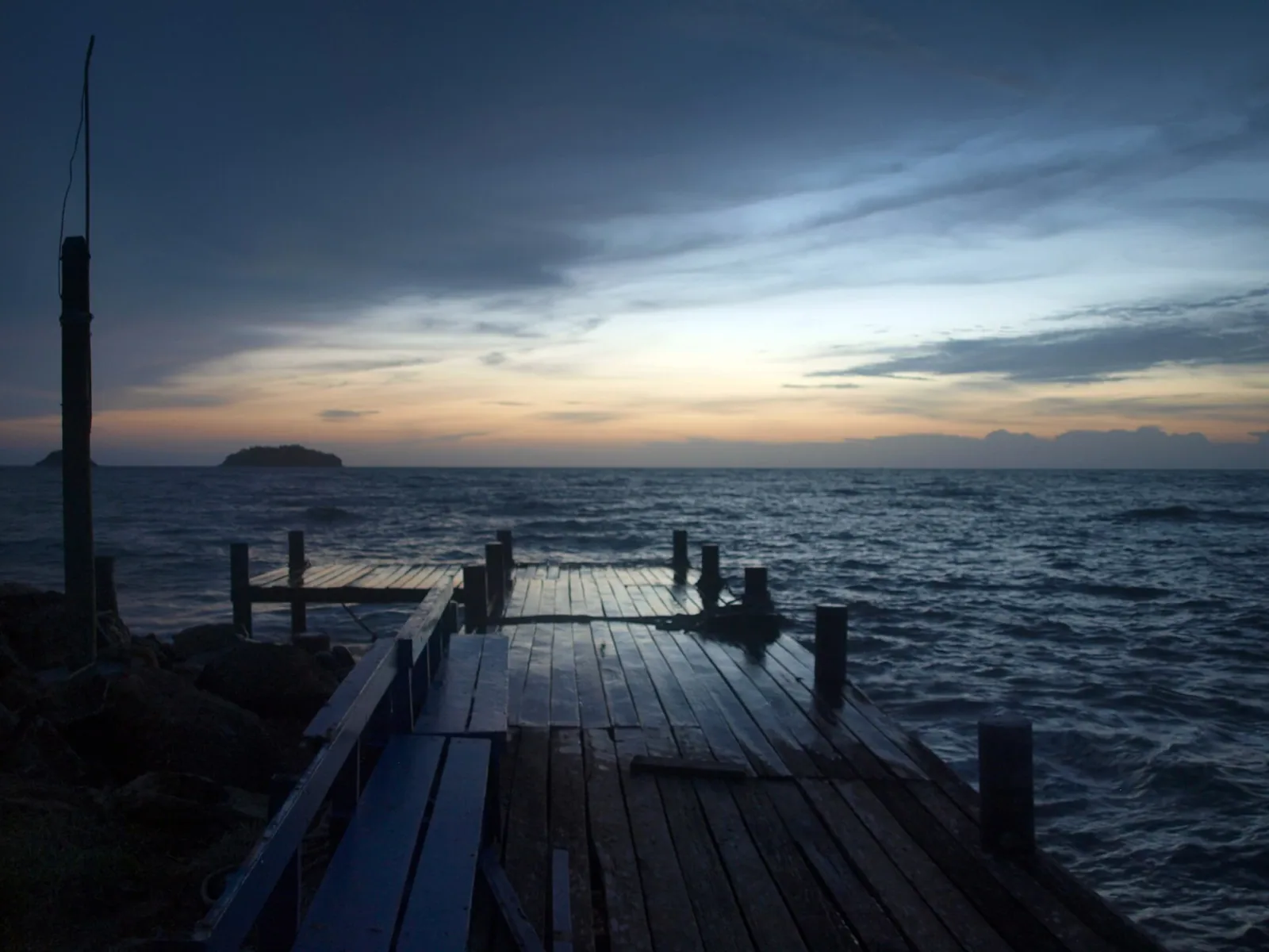 A long wooden pier stretches into the dark, wavy ocean under a dramatic sky with hues of orange and blue, with a small, dark island silhouetted on the horizon.