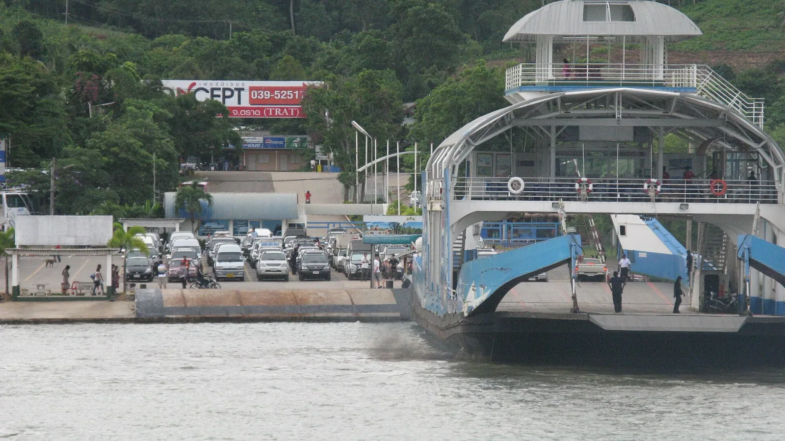 A large blue and white car ferry docked at a pier, with cars waiting to board and a forested hill in the background. A sign in the background reads 'TRAT'.