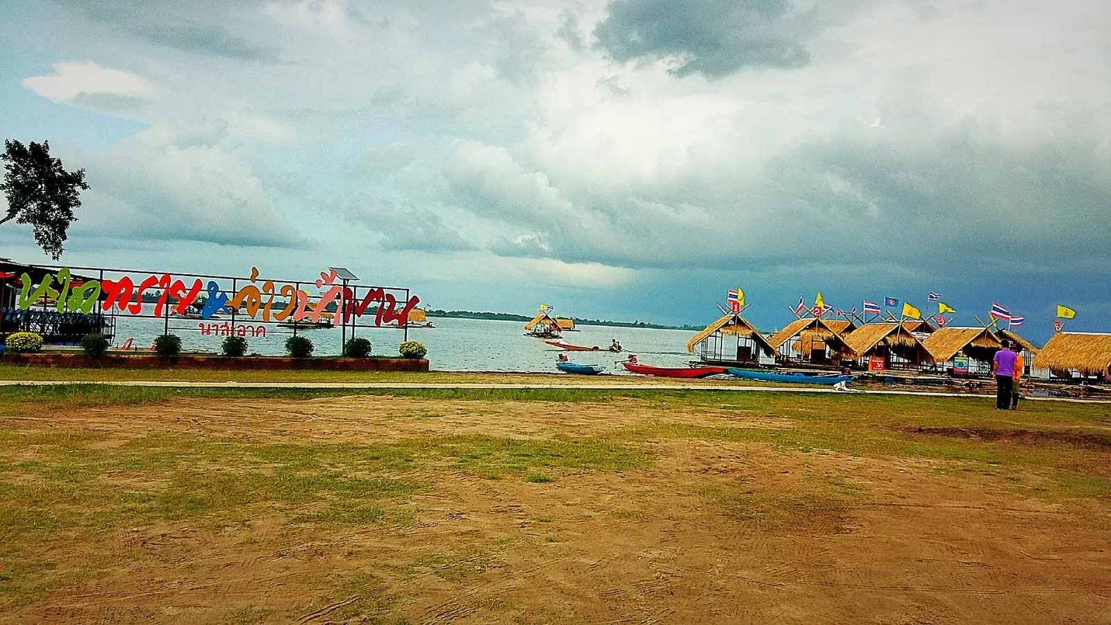 A wide shot of Ang Nam Phan reservoir in Udon Thani, Thailand, showing several floating thatched-roof restaurants decorated with flags, small boats on the water, and a colorful sign with Thai text along the shore under a cloudy sky.