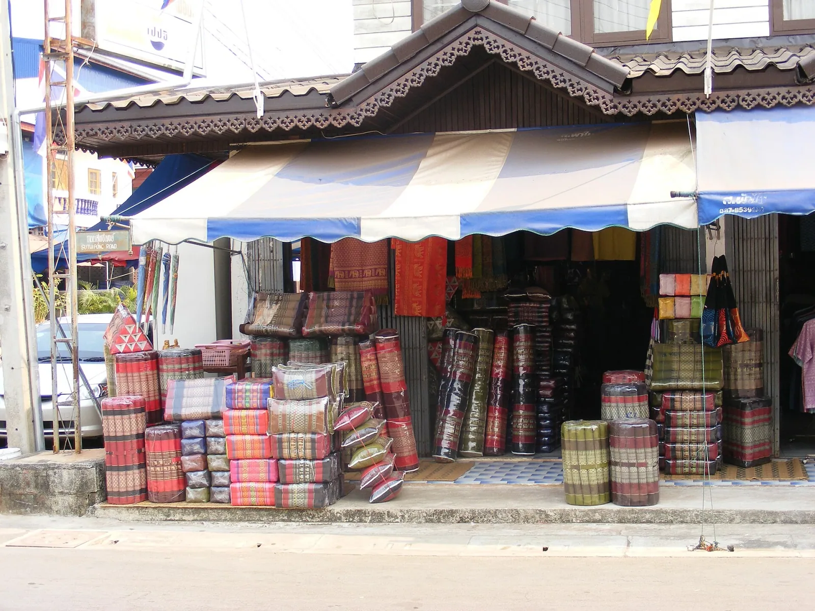 A street-side souvenir shop in Thailand with a blue and white awning, displaying stacks of colorful traditional Thai cushions and rolled mattresses.
