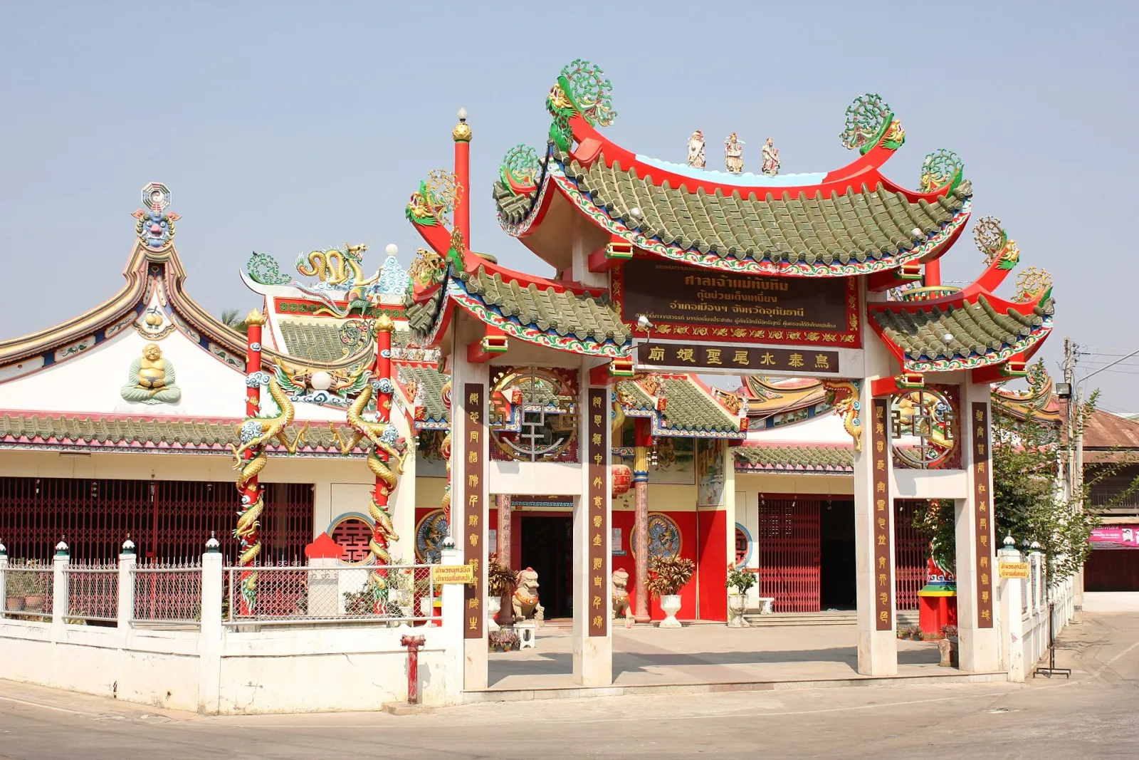 A colorful Chinese temple entrance in Uthai Thani, featuring a prominent gate with green tiled roofs, red pillars adorned with golden dragons, and a main building behind it.