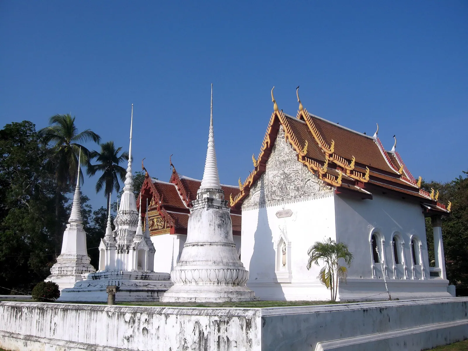 Wat Ubosatharam, a white Thai Buddhist temple with a traditional red and gold roof, stands alongside several white chedis under a clear blue sky in Uthai Thani.