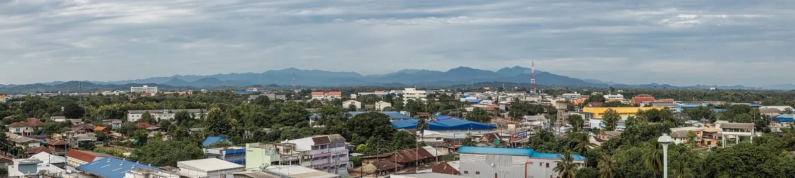 Panoramic view of Uttaradit city, showing a dense urban area with many green trees and distant mountains under a cloudy sky.