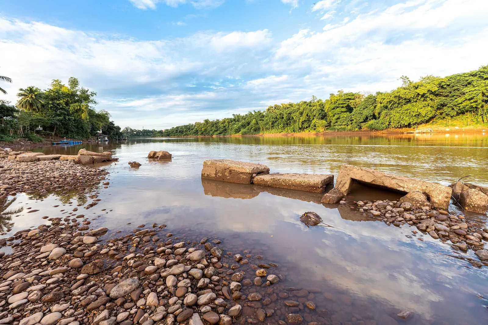 The Nan River flows under a blue sky with scattered clouds, flanked by green trees. The foreground shows a rocky riverbank with some submerged concrete blocks.