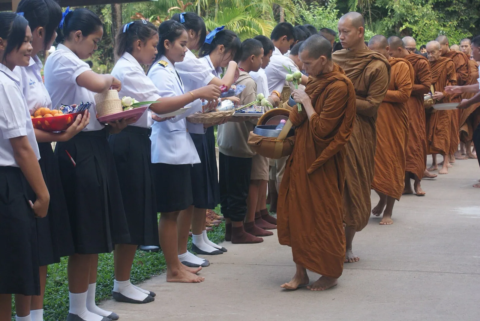 Buddhist monks in saffron robes receive alms from students in white shirts and dark skirts, who offer food and lotus flowers during a morning ceremony.