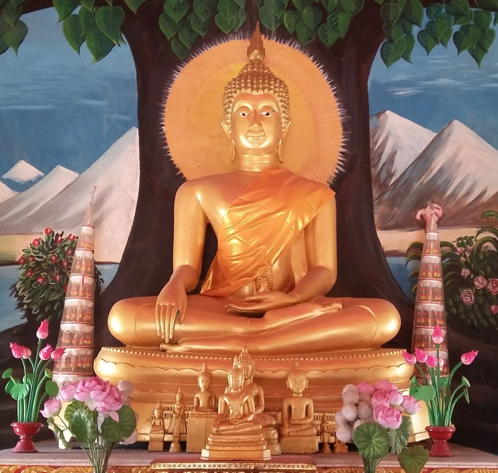 A golden Buddha statue in meditation pose, adorned with an orange robe, sits against a painted backdrop of a tree and mountains inside Wat Triswannaram in Yasothon province.